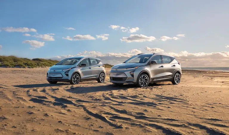 Two electric cars parked on a sandy beach under a clear blue sky.