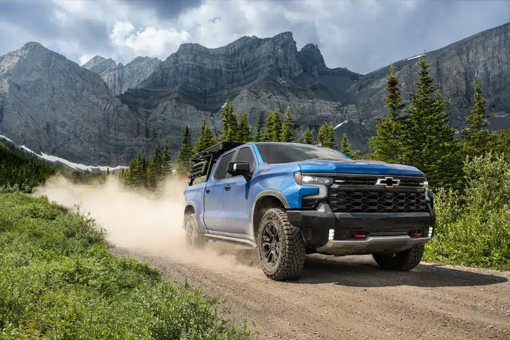 Blue truck driving on a rugged dirt road with mountains and pine trees in the background.