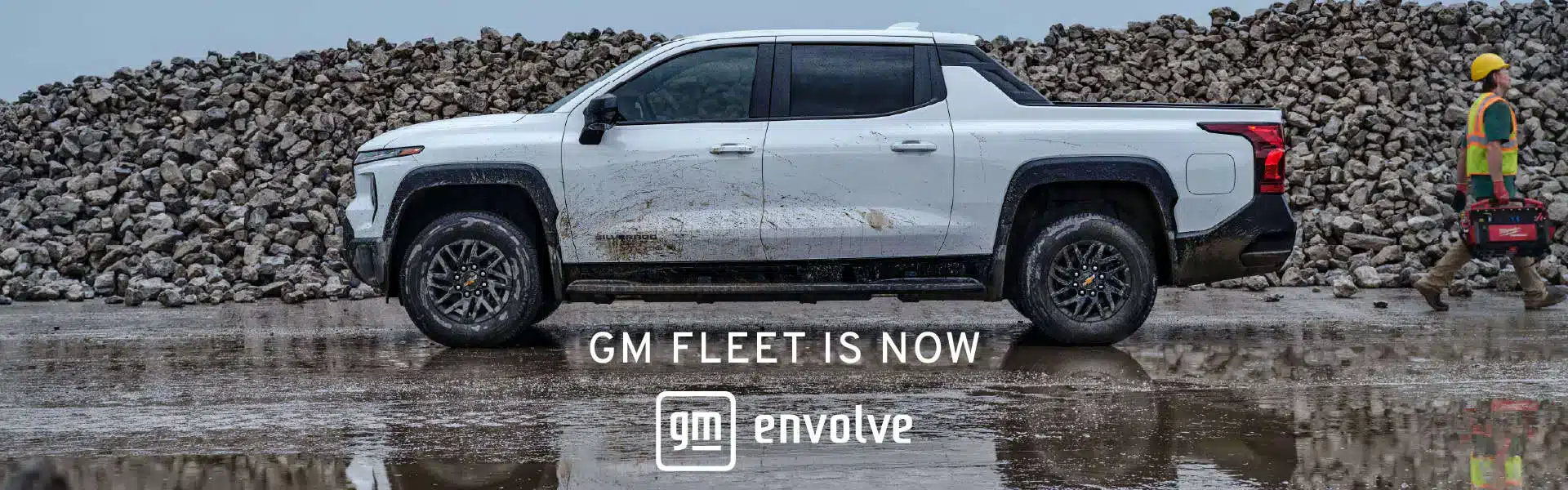 White pickup truck parked on a construction site with a worker carrying tools, featuring a backdrop of a rock pile. Text overlay reads "GM Fleet is Now Envolve."