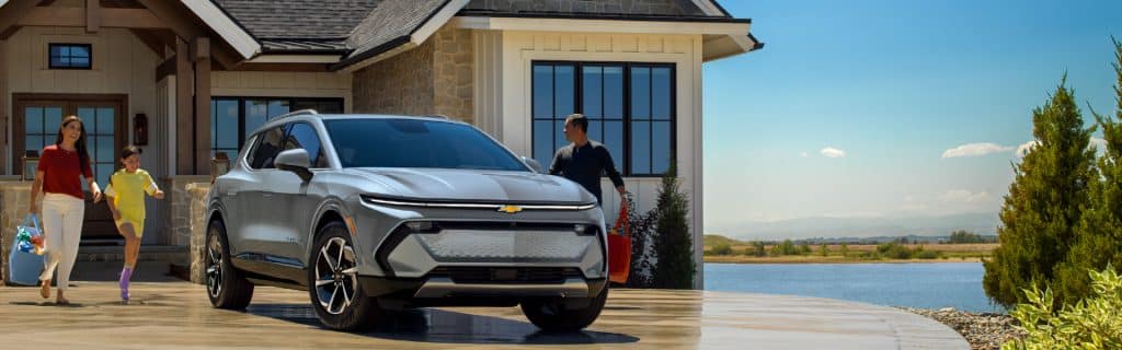 A family walks towards a modern electric SUV parked in front of a lakeside house under a clear blue sky.