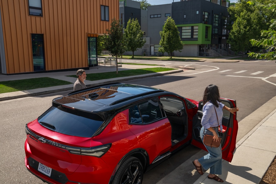 Woman entering a red SUV on a residential street with modern buildings and trees in the background.