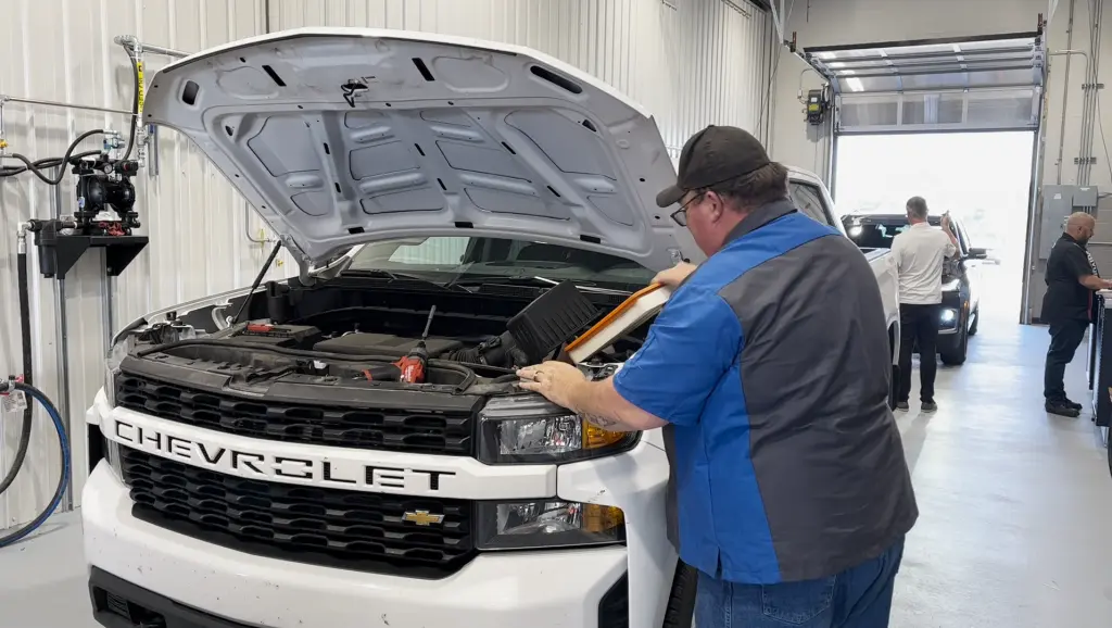 Mechanic in a blue shirt servicing a white Chevrolet truck in a garage with the hood open.