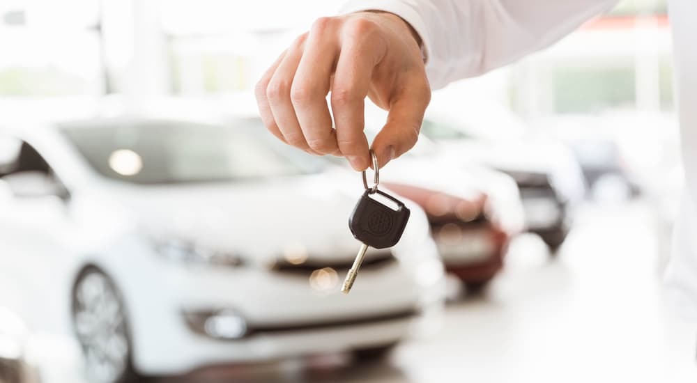 Hand holding car keys in a dealership, blurred cars in the background.