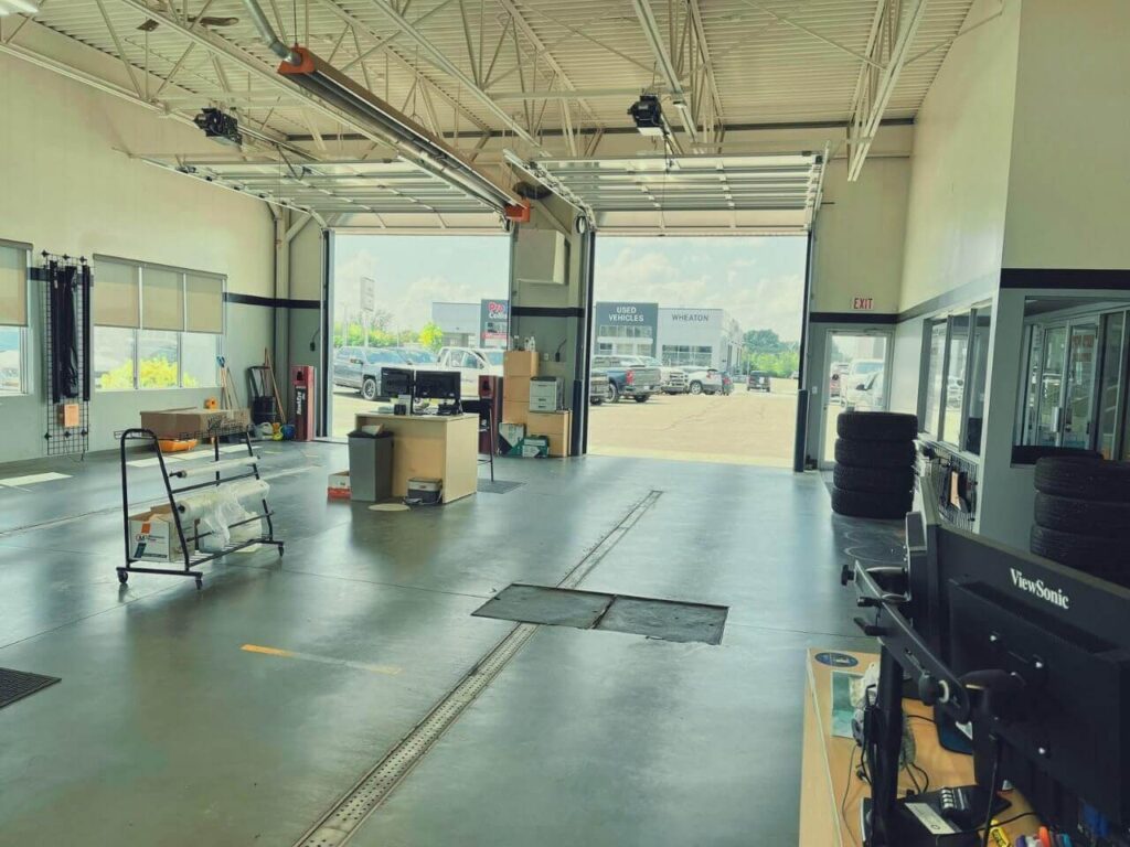 Inside a spacious car workshop with a computer station, rolled materials, and open garage doors leading outside.