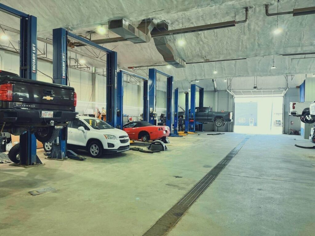 Auto repair shop interior with multiple cars on lifts, including a black truck and white car. Bright lighting.