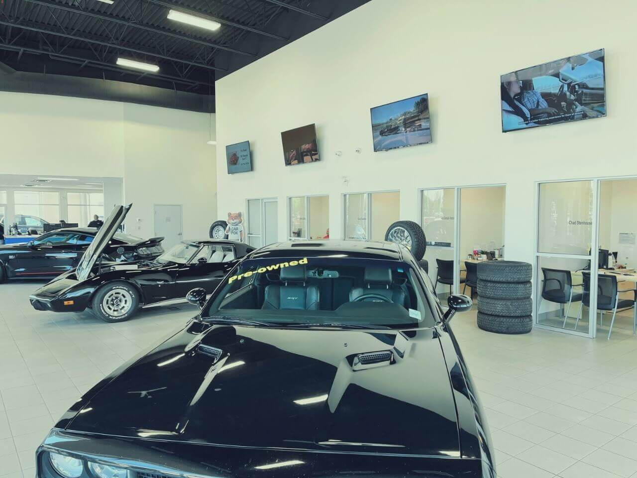 Car dealership showroom with pre-owned cars, including a black Dodge, surrounded by office spaces and tires.