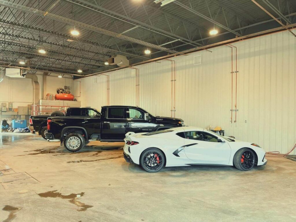 White sports car and black pickup truck parked in a spacious garage with industrial lighting.